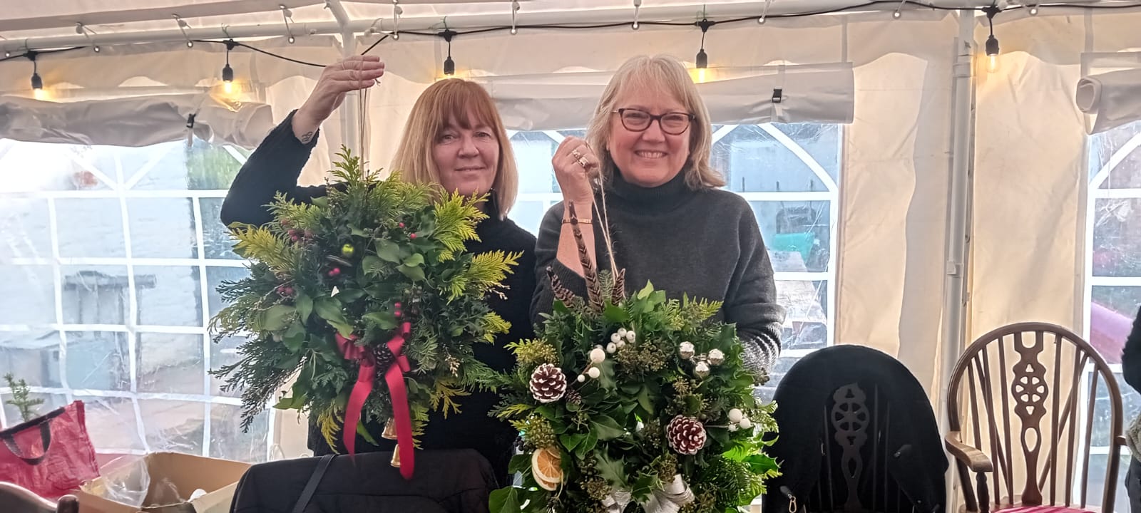 Two participants holding finished Christmas wreaths in the festive-lit marquee